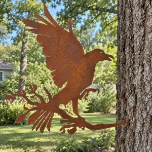 Rust-finish steel crow with wings raised on a leafy branch, its spike set into a tree trunk in a backyard